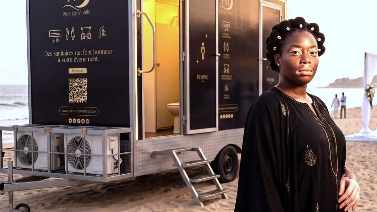 Une invitée en boubou noir brodé devant une remorque Prestige Mobile installée sur une plage au coucher du soleil, une arche de mariage dressée en arrière-plan.